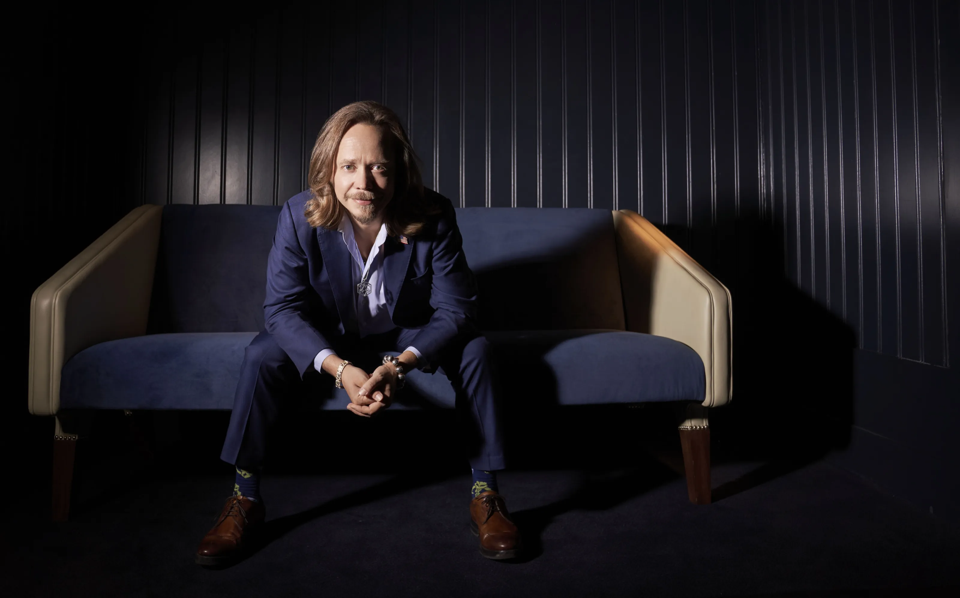 Brock Pierce seated on a navy sofa in a dark panelled room, lit by a single warm light from the upper left